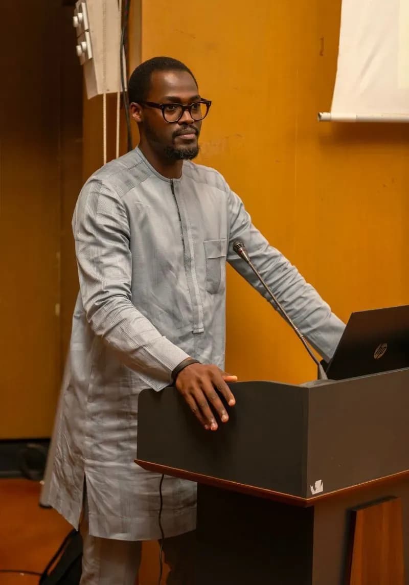 A speaker in grey traditional dress addresses the audience from the lectern